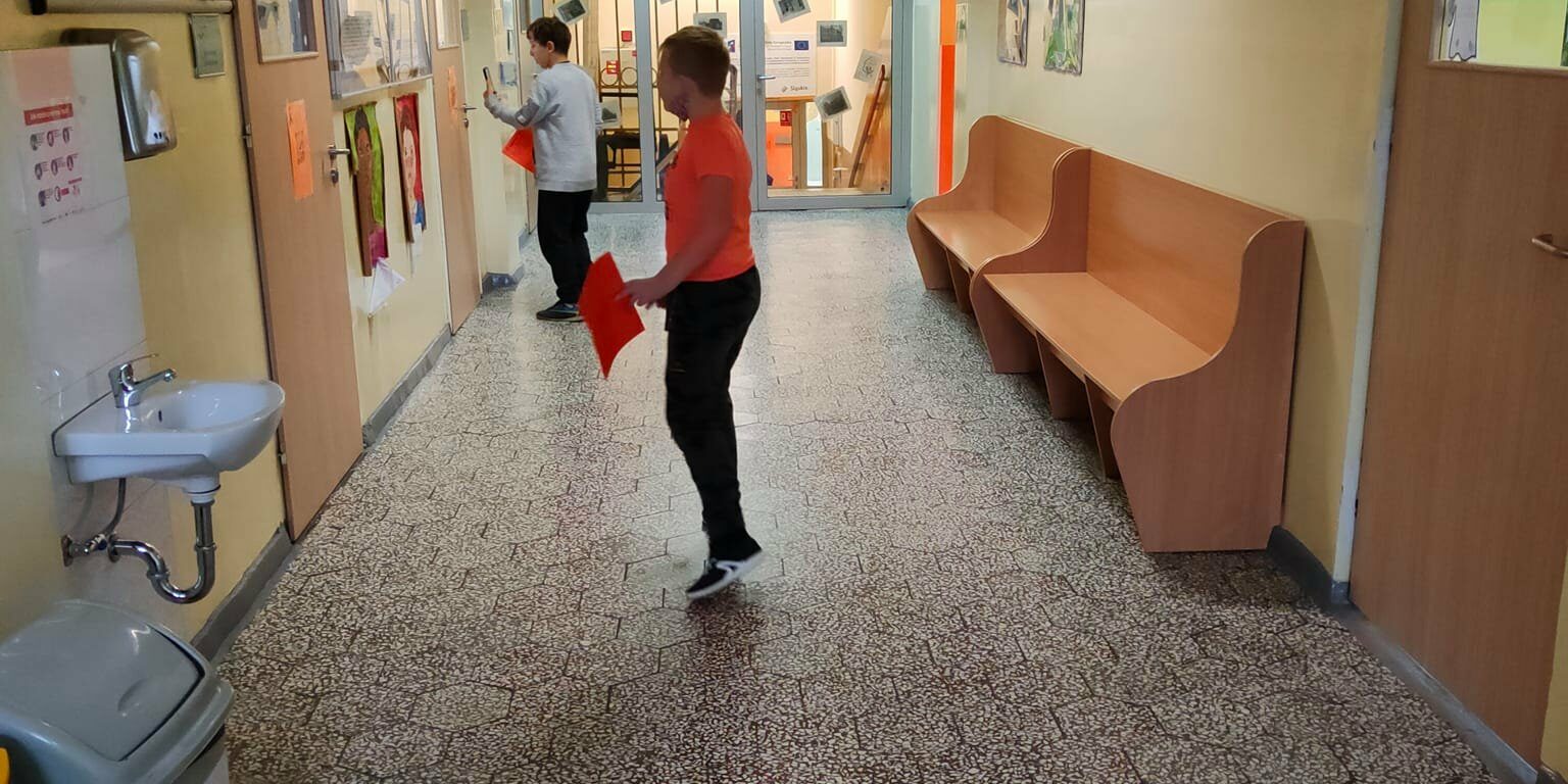 Dwoje uczniów na korytarzu szkolnym czyta teksty zawieszone na drzwiach poszczególnych klas