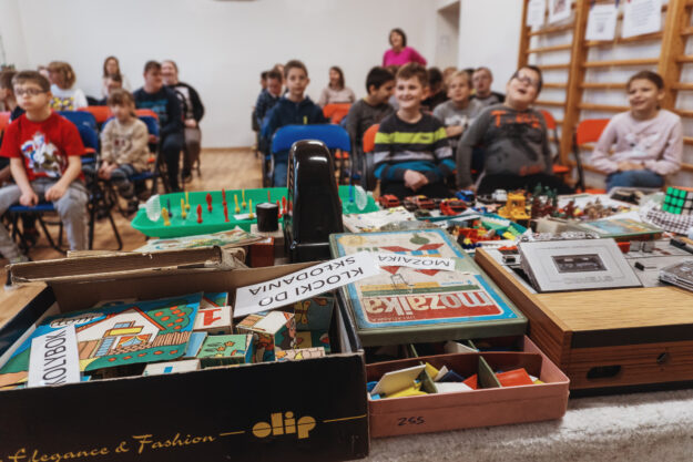 zabawki ustawione na stoliku, a w tle siedzący na krzesełkach uczniowie