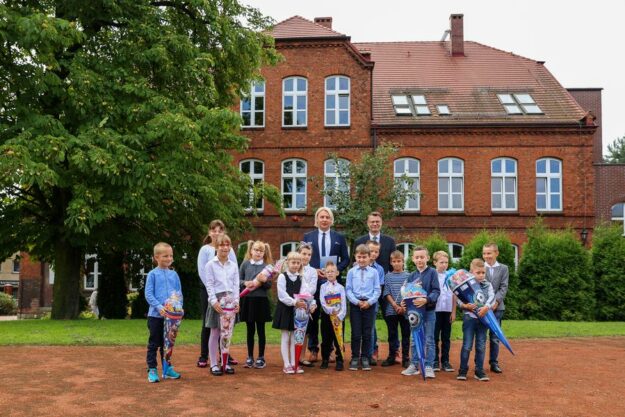 nowi uczniowie szkoły stoją razem z dyrektorami szkoły na tle budynku szkoły