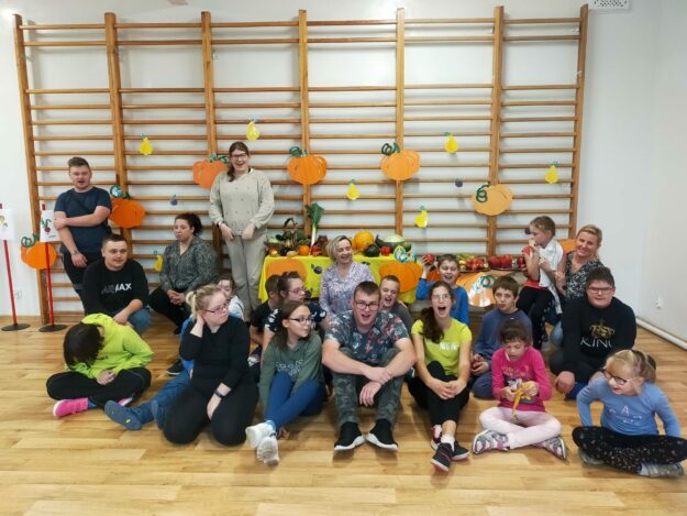 uczennicy spotkania siedzą na parkiecie a za nimi dekoracja owocowo-warzywna.