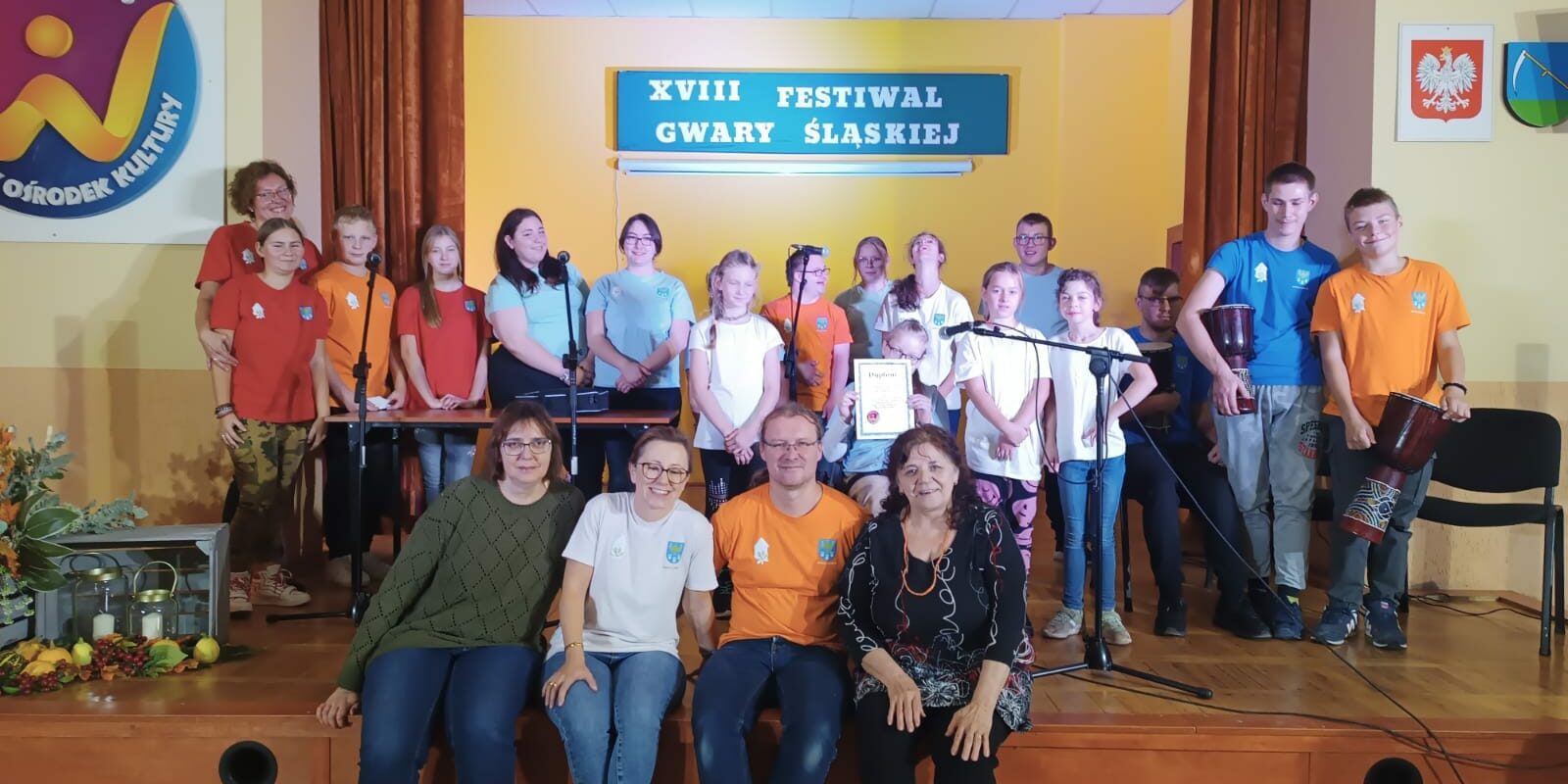 członkowie zespołu stoją na scenie, a na pierwszym planie siedzą nauczyciele wraz z członkiem jury