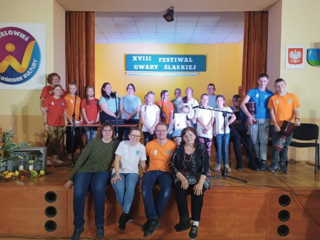 członkowie zespołu stoją na scenie, a na pierwszym planie siedzą nauczyciele wraz z członkiem jury